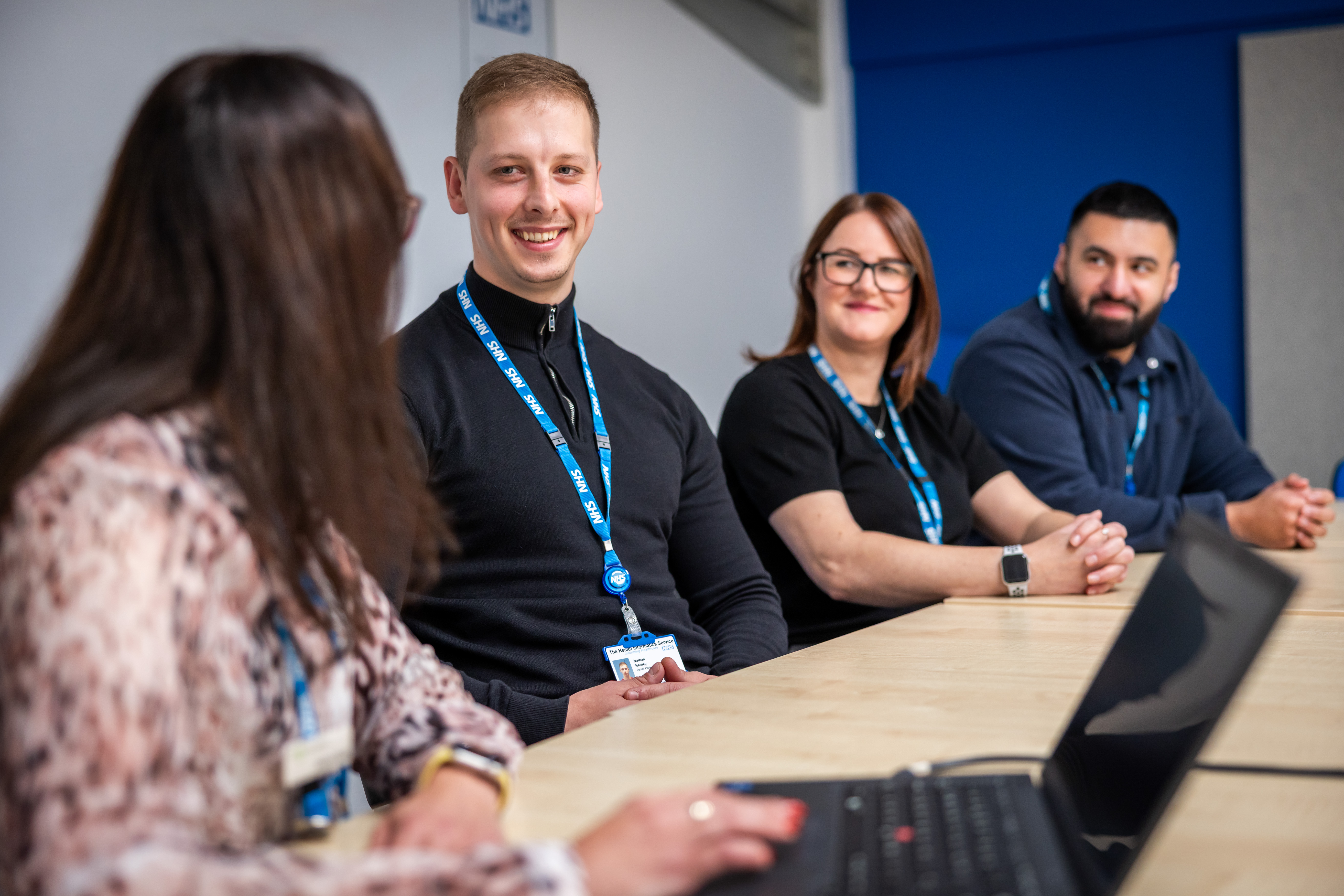 Four colleagues sit around a conference table during a meeting, wearing lanyards and smiling as they listen to one person speaking. A laptop is open on the table, and the group appears engaged in discussion.