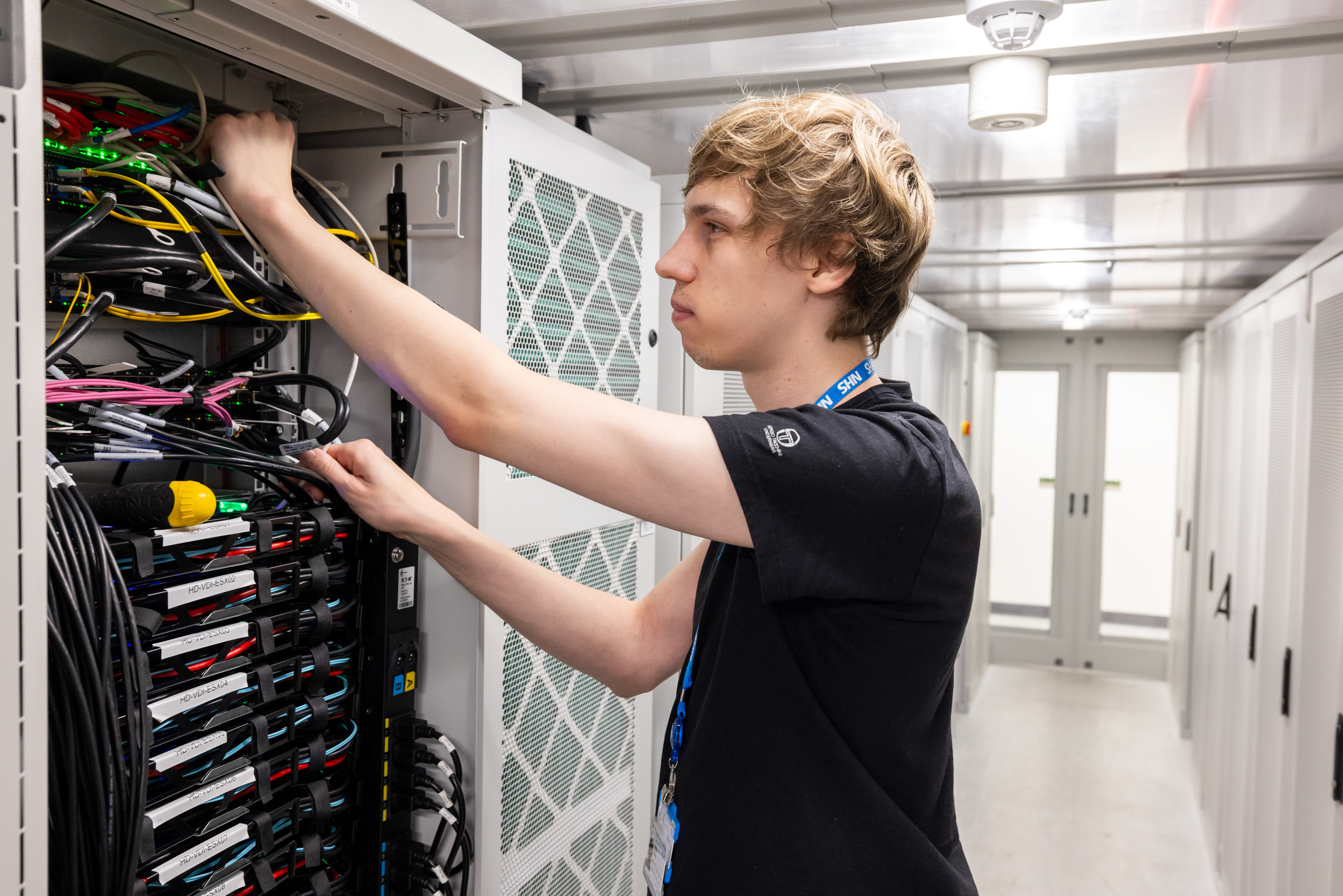 A young technician stands in a server room, reaching into an open network rack to adjust cables and equipment. Rows of servers and neatly bundled wires fill the cabinet, with more server racks visible in the background/