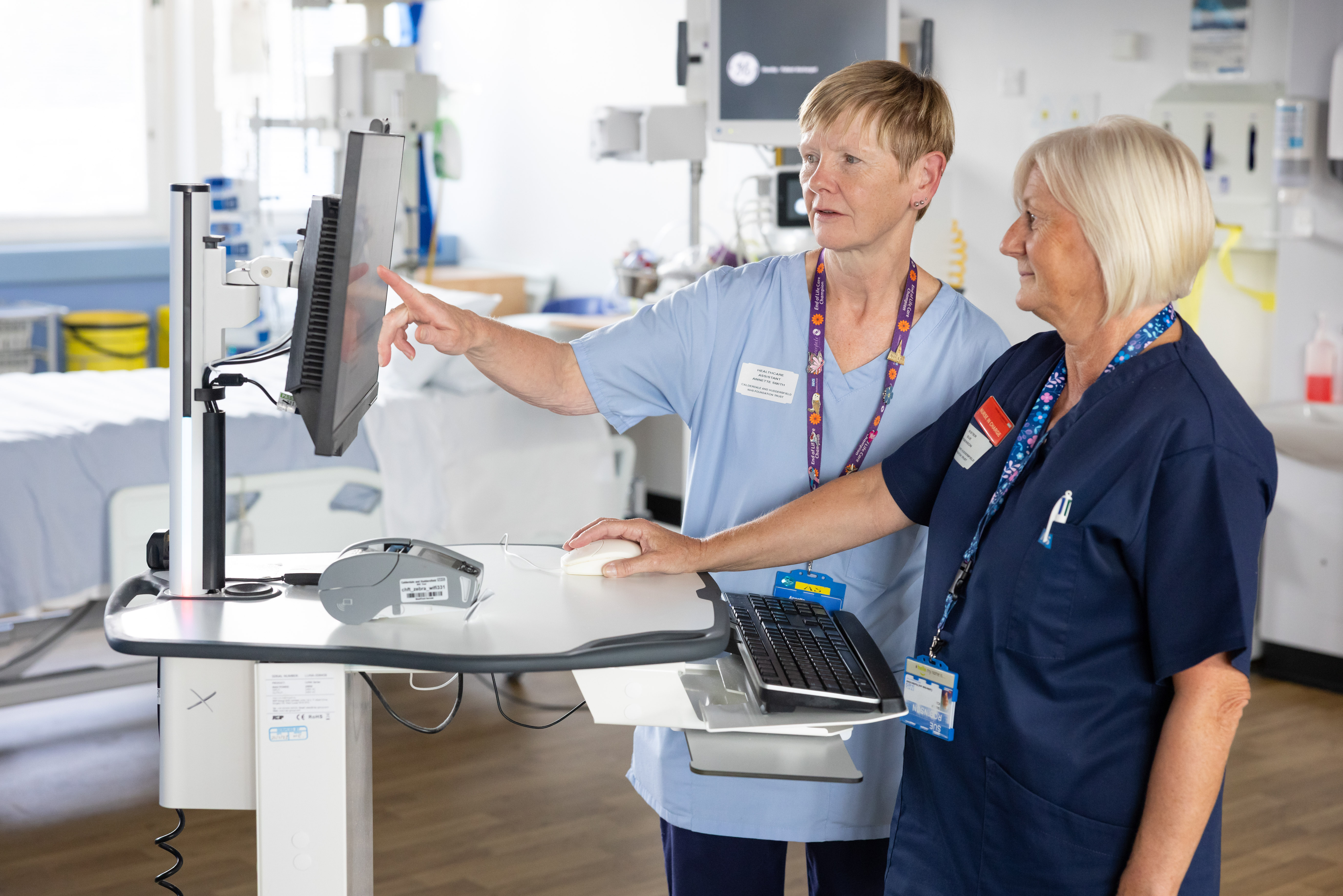 Two nurses in a hospital room stand at a mobile computer workstation. One nurse points at the monitor while the other uses the mouse, as they review patient information beside a hospital bed and medical equipment.