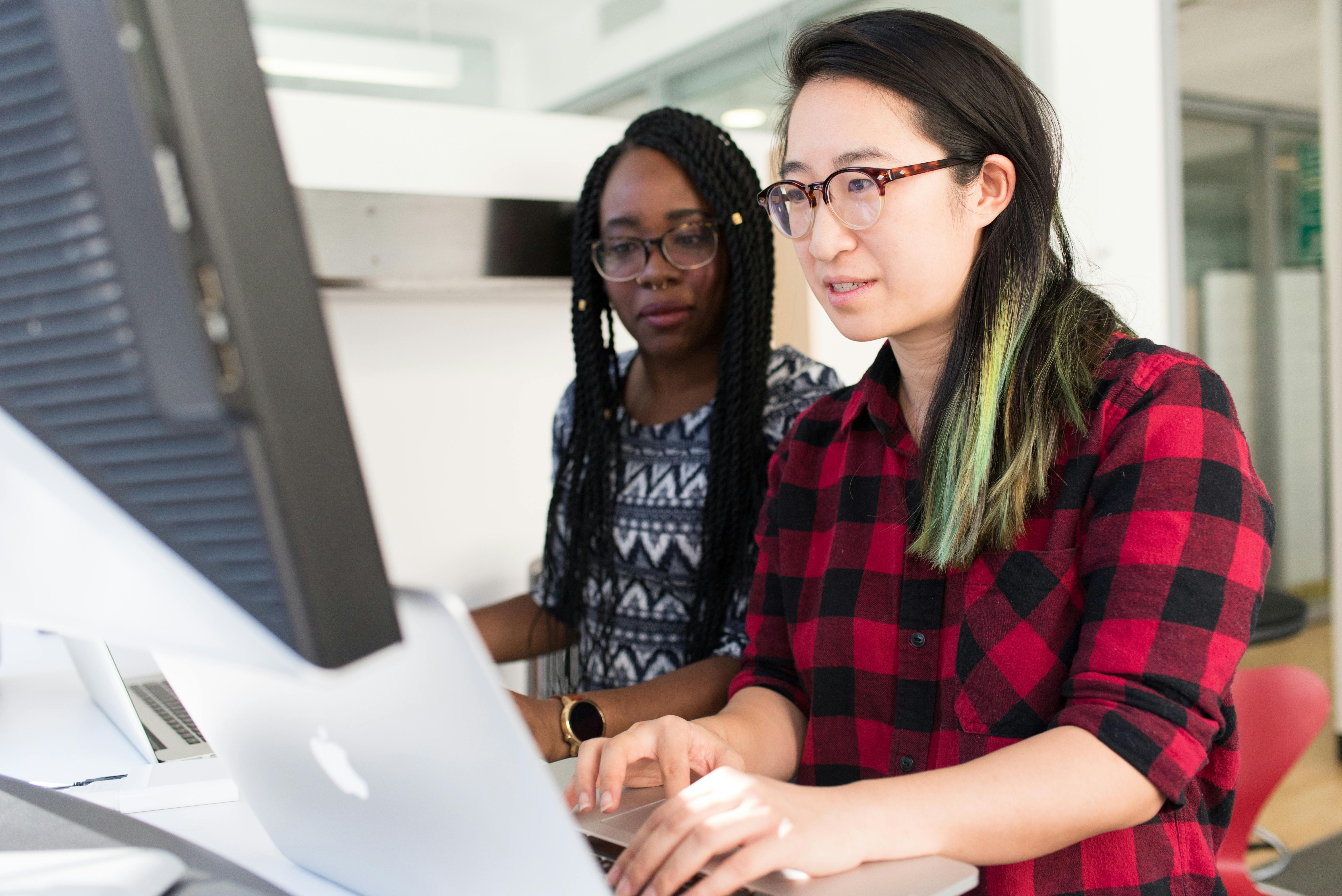 two people in an office sitting next to each other and looking at 3 different computers
