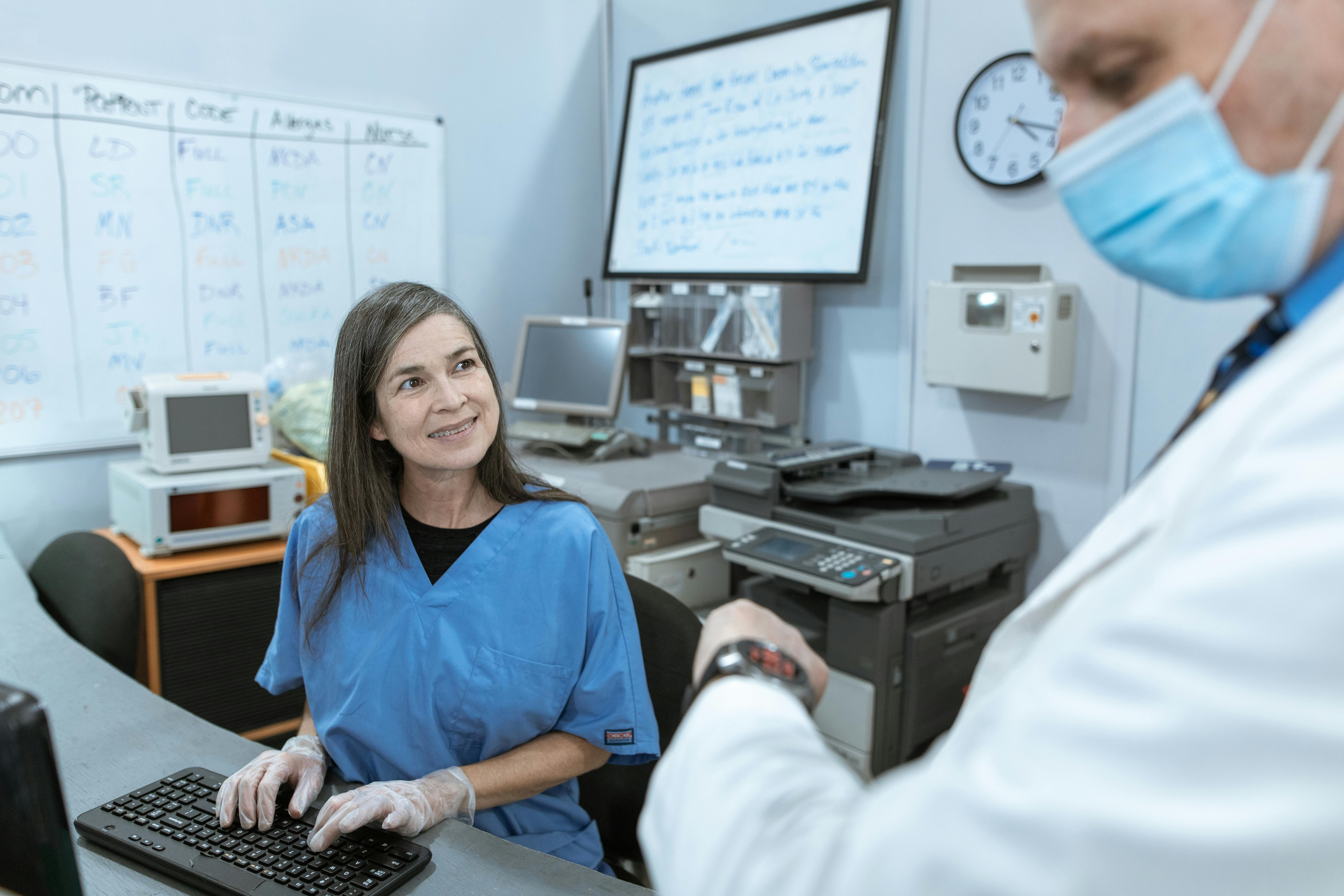 a hospital receptionist sitting in front of a computer smiling at a doctor whose wearing a mask and looking at his watch
